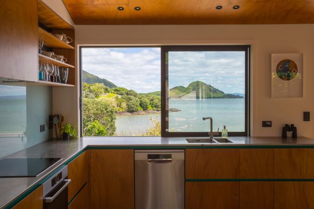 A compact, highly finished kitchen with warm timber ceiling and cabinetry, dark stone benchtops, integrated appliances, open shelving, and a large corner window overlooking a calm coastal inlet and green hills. The space has a tailored mid-century-inspired feel with strong indoor–outdoor connection and abundant natural light.