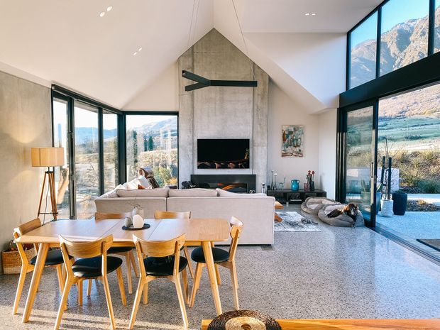 A bright, open-plan living and dining area with a dramatic pitched ceiling, floor-to-ceiling glazing, and expansive mountain views. The room features a large concrete fireplace wall with integrated shelving, a light timber dining table and chairs, a neutral sectional sofa, polished concrete flooring, and contemporary black-framed windows and doors.
