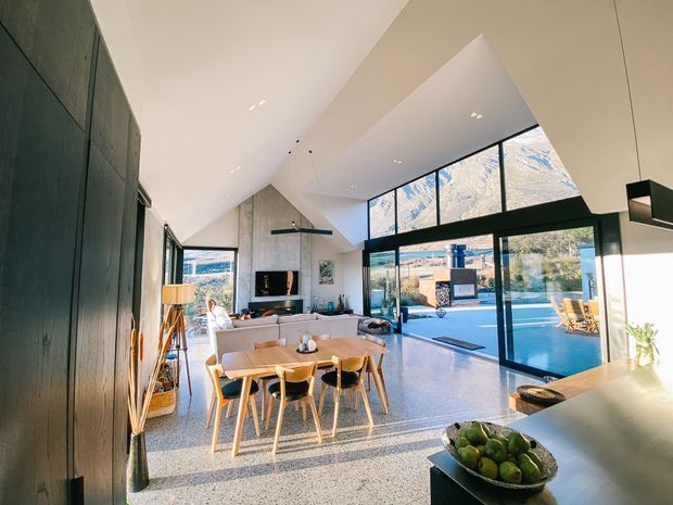 Open-plan contemporary living and dining area with a dramatic vaulted ceiling, expansive black-framed glazing, polished terrazzo flooring, and direct views to a swimming pool and landscaped outdoor area. A dark timber-like built-in wall, fireplace, dining table, kitchen island, and relaxed neutral furnishings create a bright, refined interior.
