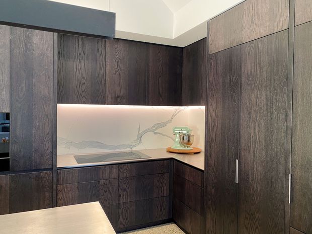 A compact contemporary kitchen with dark, vertically grained timber cabinetry wrapping around two walls. The corner features a pale veined stone splashback with integrated warm LED lighting, a sleek cooktop, and a small appliance area. Tall pantry-style cabinets occupy the right side, while a light stone island or benchtop is visible in the foreground.