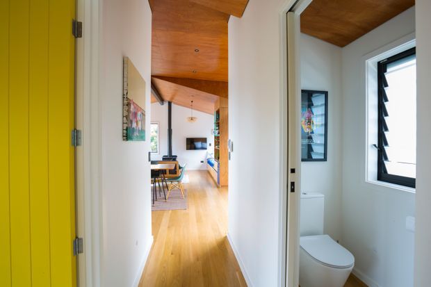 Bright interior view down a narrow hallway with warm timber flooring and a timber-lined ceiling. A dining area and living space are visible at the end, while an open bathroom with a white toilet and black-framed awning window is visible on the right. White walls, colourful artwork, and strong yellow door trim add contrast.