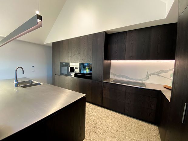Contemporary kitchen featuring a large island with integrated sink, dark timber cabinetry, black appliances, and a white marble-look splashback illuminated by under-cabinet lighting. A light terrazzo-style floor and sculptural linear ceiling light add contrast to the refined, minimalist space.