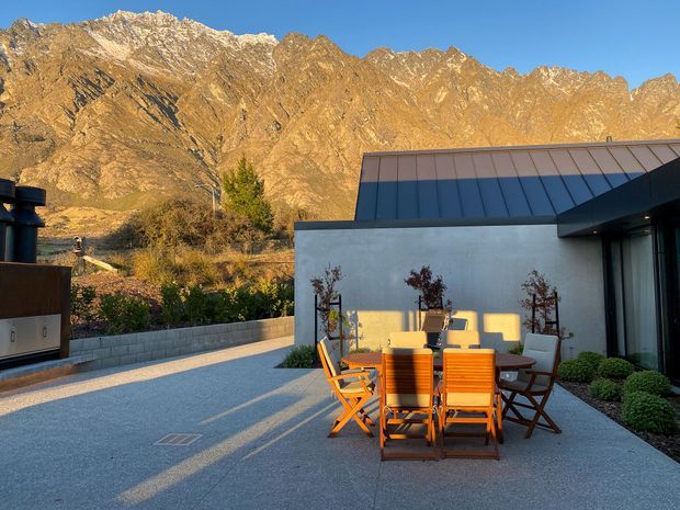 A modern single-storey home or pavilion with a low-pitched metal roof and smooth light-coloured rendered walls, opening onto a spacious paved outdoor dining terrace. Timber outdoor dining chairs and a table sit in warm late-afternoon sunlight, with planted borders and a low masonry wall. Dramatic rugged alpine mountains rise immediately behind the property.