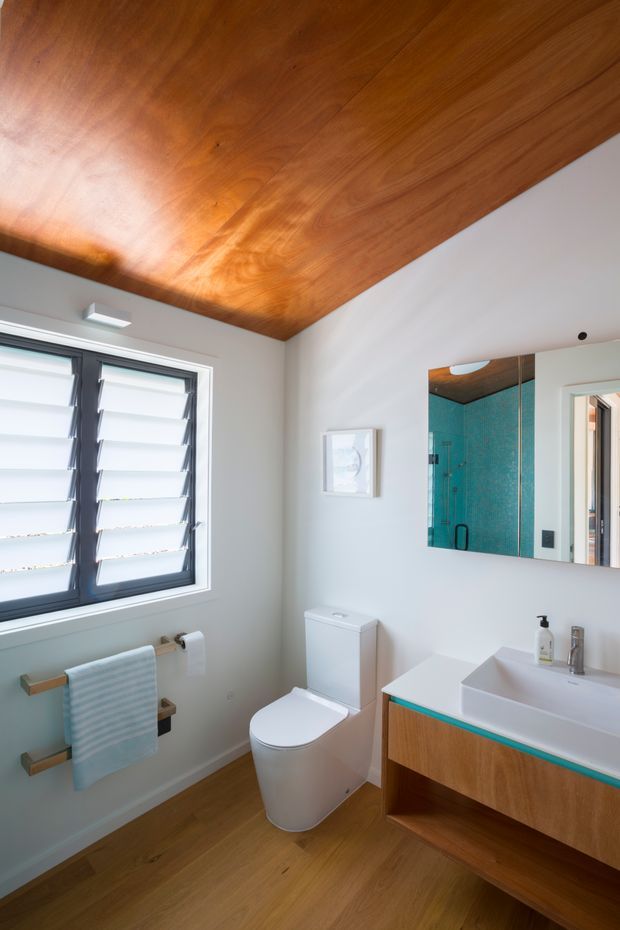 Bright contemporary bathroom with a wall-mounted timber vanity, white rectangular basin, chrome tapware, white toilet, louvred window, timber flooring, and a striking warm timber-lined ceiling. A large mirror reflects a turquoise tiled shower area.
