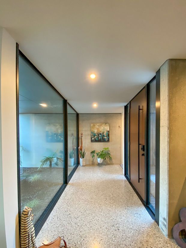 A long, contemporary interior hallway with polished terrazzo flooring, white ceiling, recessed downlights, and full-height dark-framed glass partitions on the left enclosing a planted internal garden or courtyard. Timber-toned doors and dark-framed glazing line the right side, while a textured concrete or stone feature wall anchors the foreground. Potted plants and artwork add warmth at the end of the corridor.