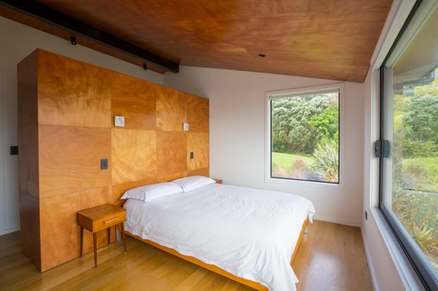 A compact, warm bedroom with a low-profile bed positioned against a wall of built-in plywood cabinetry. The room has honey-toned timber flooring and a matching timber-clad ceiling, white walls and bedding, and large windows overlooking lush greenery. A small timber bedside table sits beside the bed, while dark ceiling-mounted hardware adds a subtle industrial detail.