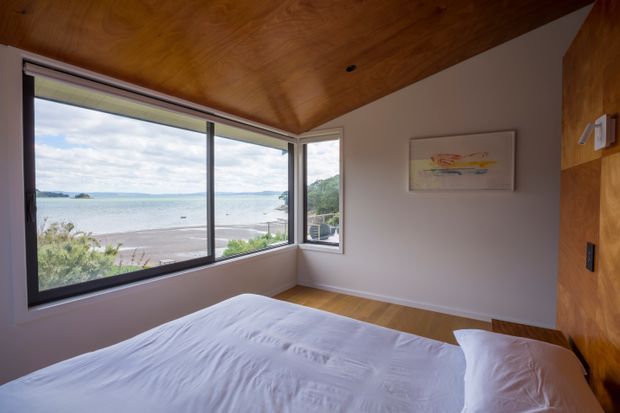 A serene bedroom with a broad corner of dark-framed glazing overlooking a sandy waterfront or beach. The room features a low-profile bed with white linen, warm timber flooring, a rich timber-lined ceiling and matching timber wall panel, white walls, and a minimal framed artwork. The expansive view and natural materials create a calm, coastal atmosphere.