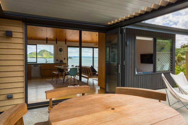 A covered outdoor entertaining area with a timber dining table and bench seating opens directly into a compact open-plan kitchen and living space. The interior features warm timber walls and cabinetry, dark-framed glazing, and a pitched or angled white-lined ceiling. Large sliding openings connect the spaces, with a bright coastal landscape and water visible beyond.