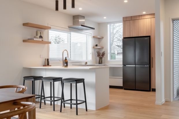 Bright contemporary kitchen with a white island and integrated seating, light timber flooring, white walls, open timber shelves, stainless-steel rangehood, and a tall dark refrigerator set within full-height timber cabinetry. Large windows with blinds provide natural light, and part of a dining area is visible in the foreground.