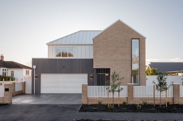 Contemporary multi-level residential house viewed from the street, featuring a prominent warm brick gabled facade, tall narrow window, dark vertical cladding around the integrated garage, a pale metal-roofed side volume, and a low brick-and-slat boundary fence with young landscaped trees.