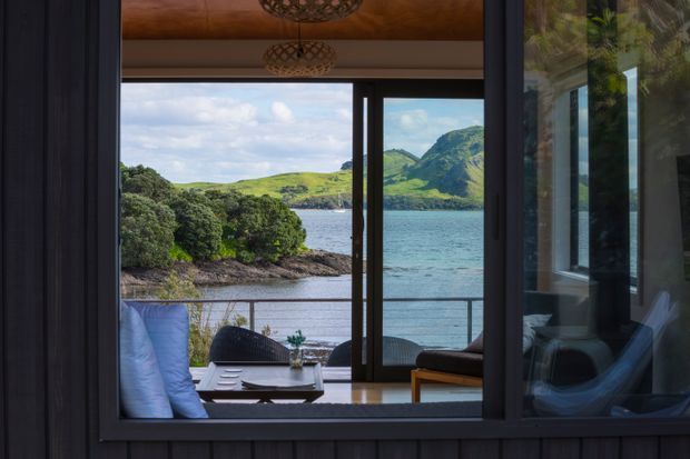 View from inside a dark-framed, glazed room toward a calm coastal inlet, with a covered deck furnished with outdoor seating and a low table. The scene includes timber-lined soffit detailing, expansive window and sliding-door framing, lush shoreline vegetation, and green hills across the water.