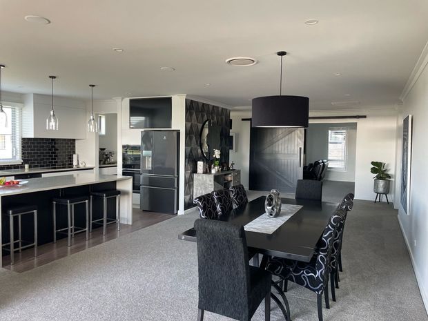 Open-plan contemporary kitchen, dining and living area with a monochrome palette. The kitchen features dark cabinetry, a white benchtop, black subway-tile splashback, pendant lights and integrated stainless-steel appliances. A dark dining table with patterned upholstered chairs sits in the foreground, while the living zone includes a large black pendant, textured dark feature wall, fireplace or media joinery, and neutral carpet.