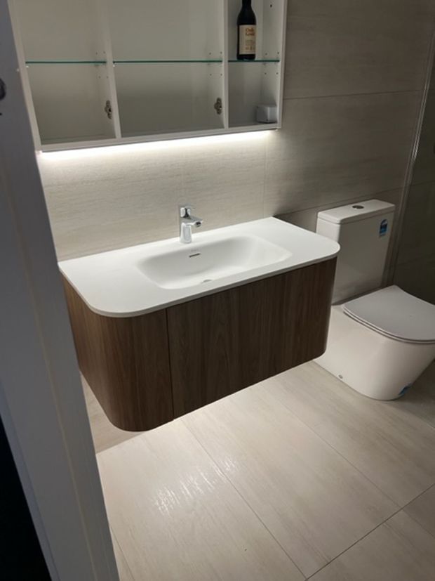 Compact contemporary bathroom with a wall-hung wood-look vanity, integrated white basin and chrome tapware. Large light beige floor and wall tiles create a clean, warm-neutral finish, while mirrored wall cabinetry with open glass shelving and under-cabinet lighting adds storage and illumination. A modern white toilet sits beside the vanity.