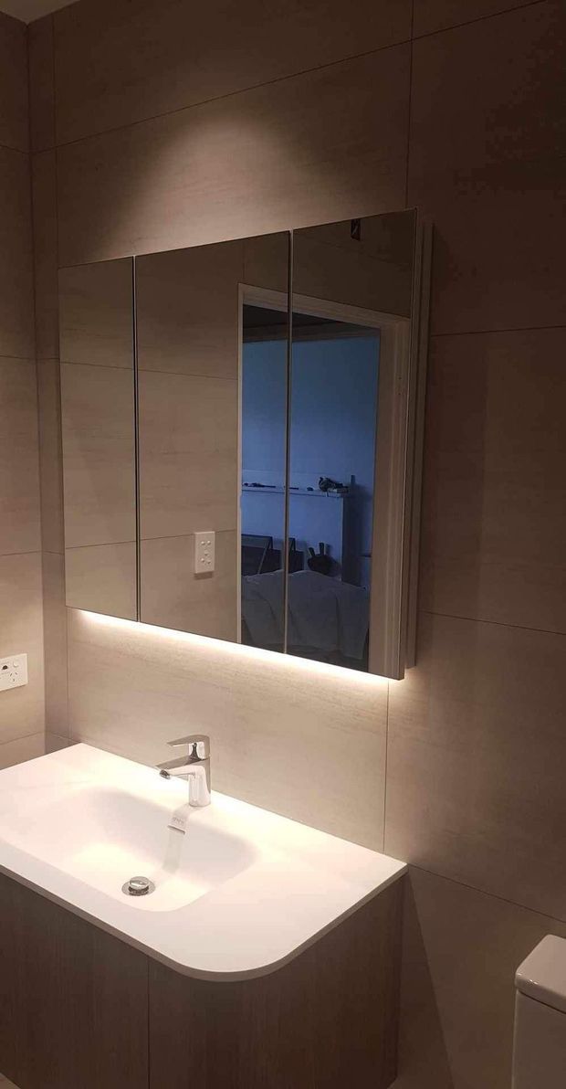 A compact contemporary bathroom with large taupe-beige wall tiles, a white integrated vanity basin with a chrome tap, dark wood-toned cabinetry, and a mirrored wall cabinet with under-cabinet LED lighting. A toilet is partially visible at the right edge, and the mirror reflects an adjacent bedroom or room.