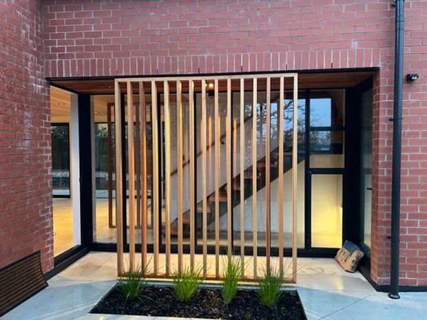 Exterior view of a contemporary brick building entrance or courtyard opening, with black-framed glazing and doors behind a vertical timber batten screen. A timber staircase is visible through the glazing, with pale paving and a narrow landscaped planter in the foreground.