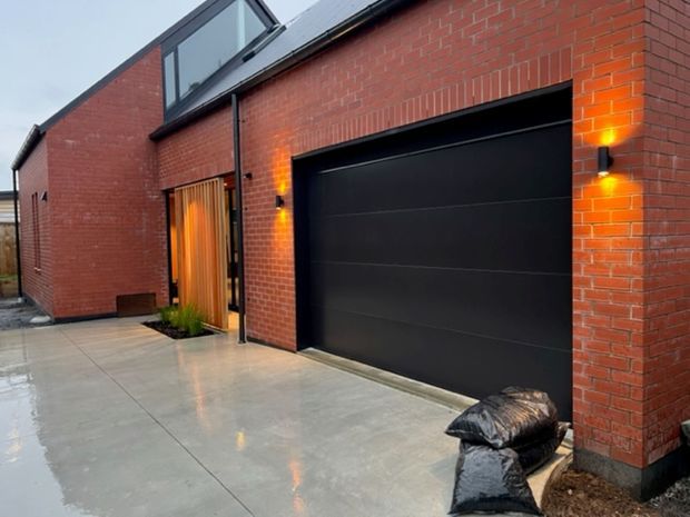 Exterior view of a contemporary red-brick building with a large matte-black sectional garage door, black-framed glazing, a timber entry door, warm wall sconces, and a broad smooth concrete driveway. The simple angular roofline and exposed brick facade create a clean modern industrial character.