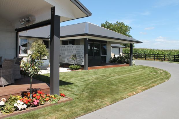 Single-storey contemporary residence viewed from the landscaped front yard, featuring a low-pitched skillion-style roof, dark structural posts and window frames, light rendered walls, a covered outdoor deck, paved driveway, and manicured lawn with garden beds. Rural fields are visible beyond the property.