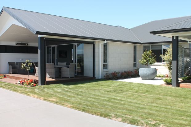 Single-storey contemporary residential house viewed from the front, with a light rendered or masonry facade, grey metal pitched roof, covered outdoor entertaining areas, dark-framed glazing, manicured lawn and landscaped garden beds. The central covered patio opens to an indoor living area, while a second covered outdoor space is visible on the right.