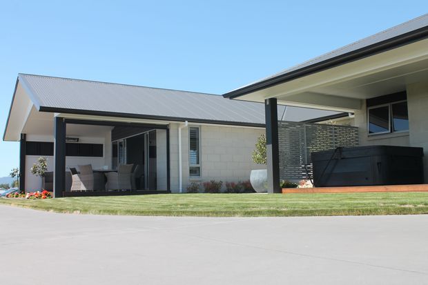 Contemporary single-storey home exterior with a broad pitched metal roof, pale masonry walls, dark structural posts and window frames, and a covered outdoor living area. The view shows a landscaped lawn, paved driveway, outdoor seating, and a partially screened terrace with a large dark spa or outdoor fixture. Bright, clean suburban residential setting.