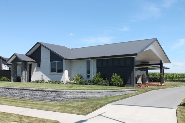 Single-storey contemporary suburban house viewed from the street, with a broad dark grey gabled roof, light rendered or masonry walls, dark vertical-panel feature cladding around the garage, stone-clad entry columns, and an attached open-sided carport. The landscaped front yard has stepped dark retaining walls, lawn, shrubs, and a concrete driveway.