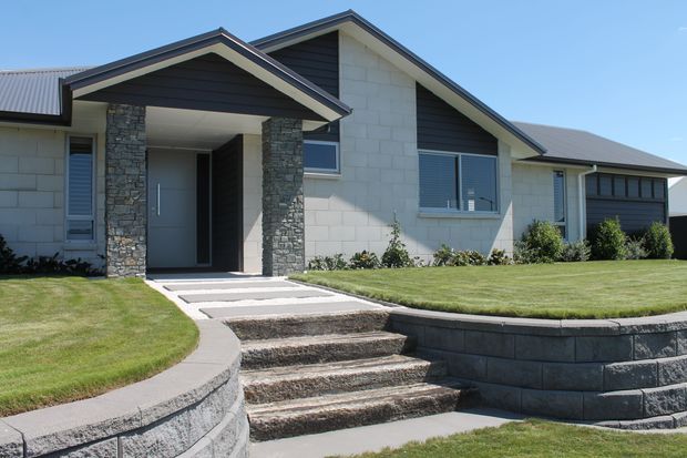 Single-storey contemporary suburban home viewed from the front, featuring a gabled roof, pale masonry block walls, dark horizontal cladding in the gable ends, stone-clad entry columns, and a recessed front door. Curved concrete pathways, stepped landscaping, retaining walls, and a neatly maintained lawn define the approach.