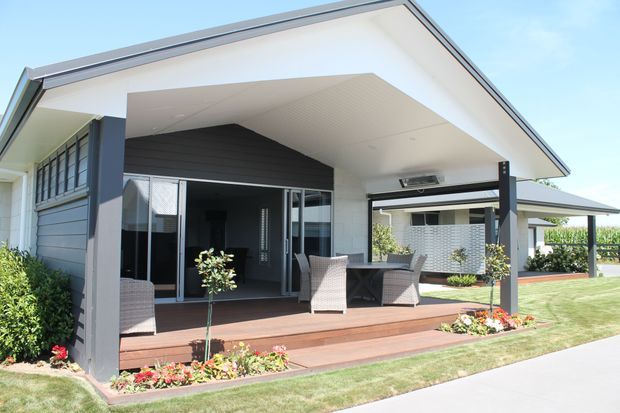 Single-storey contemporary house exterior with a broad covered patio and timber decking. The facade combines dark horizontal cladding, white rendered surfaces, large glazed openings, and a prominent skillion-style roof canopy. Outdoor dining and lounge furniture sit beneath the sheltered terrace, with landscaped garden beds and a lawn in front.