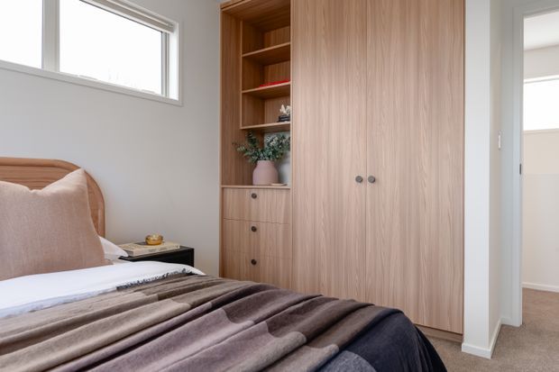 A calm, softly styled bedroom with a timber bedhead, layered neutral bedding, and integrated light-wood wardrobe and shelving joinery. A small bedside table sits beneath a high window, while an open doorway leads to an adjacent carpeted space.