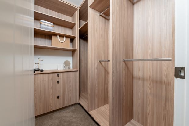 Compact walk-in wardrobe with floor-to-ceiling light wood-grain cabinetry, open shelving, hanging storage, and integrated lower cupboards. A small built-in vanity or utility counter with a tap is positioned to the left, while the room has a neutral white wall finish and dark carpet flooring. The design is clean, contemporary, and highly organized.