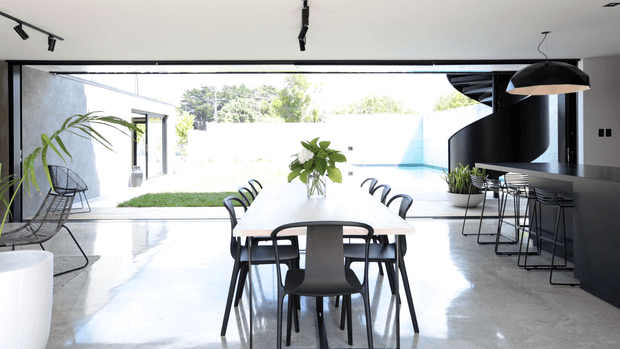 A bright, open-plan contemporary dining area with a long light timber dining table and black chairs positioned in front of fully open floor-to-ceiling glazing. A polished concrete floor extends toward a landscaped courtyard and swimming pool outside. On the right is a dark sculptural curved staircase or feature wall with a black kitchen or bar counter, stools, pendant lighting, and indoor plants. The space has a minimalist monochrome palette, strong indoor-outdoor connection, and generous natural light.