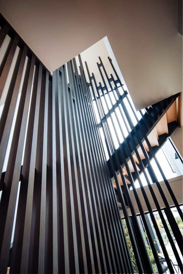 Upward-looking interior view of a tall stairway or landing enclosed by dramatic vertical dark timber and black metal screening. Floor-to-ceiling glazing and a high ceiling bring in strong daylight, while the layered slatted balustrades create a sculptural, contemporary architectural feature.
