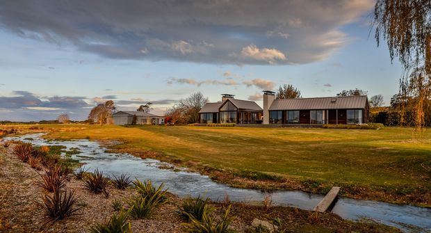 Wide exterior view of a low, single-storey rural residence beside a narrow creek or drainage channel. The home comprises several connected or closely grouped wings with dark metal pitched roofs, extensive glazing, and a restrained contemporary composition. Open lawn, wetland-like planting, and expansive flat landscape surround the building under dramatic evening cloud.