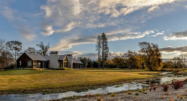 Wide exterior view of a low, contemporary residential building set within an expansive landscaped rural property. The home combines dark vertical timber-clad gabled volumes with pale rendered rectangular sections and extensive glazing. A broad lawn, shallow stream or wetland edge, mature trees, gravel landscaping, and dramatic sunset sky surround the house.
