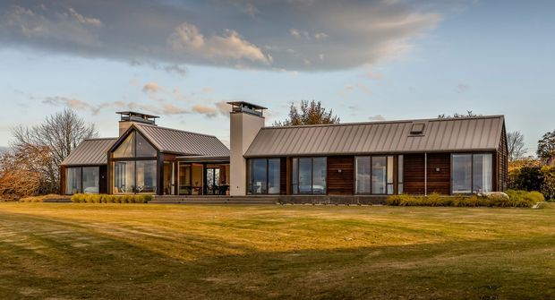 A low-profile modern residence viewed from across a broad lawn. The house has two connected wings with dark standing-seam pitched roofs, extensive floor-to-ceiling glazing, dark timber or wood-look cladding, and a prominent central masonry chimney. The surrounding landscaped grounds and autumnal trees are visible under a partly cloudy sky.