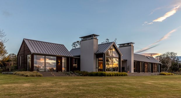 A wide exterior view of a low-rise contemporary residential home set behind an expansive lawn. The building is composed of several connected gabled volumes with dark vertical timber cladding, standing-seam metal roofs, large glazed openings, and prominent rectangular chimney or flue towers with metal caps. Warm interior lighting is visible through the glazing, and landscaped planting runs along the facade.