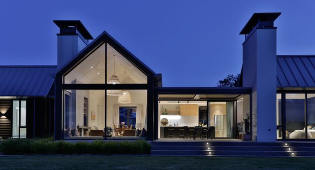 Twilight exterior view of a contemporary single-storey residence with multiple pitched roof volumes, prominent glazed gable ends, dark metal roofing and cladding, and tall chimney forms. The illuminated interiors reveal a living area and open-plan kitchen/dining space, while recessed step lighting defines the terrace edge.