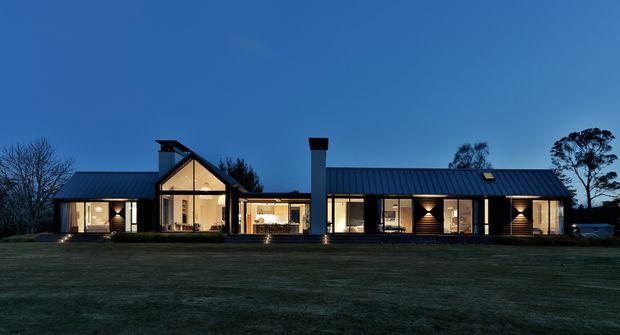 A single-storey contemporary rural residence photographed at dusk, with a long low-profile form, dark standing-seam metal pitched roofs, extensive illuminated glazing, and a prominent central double-height gabled section. The home sits behind a broad lawn with sparse trees visible around the perimeter.