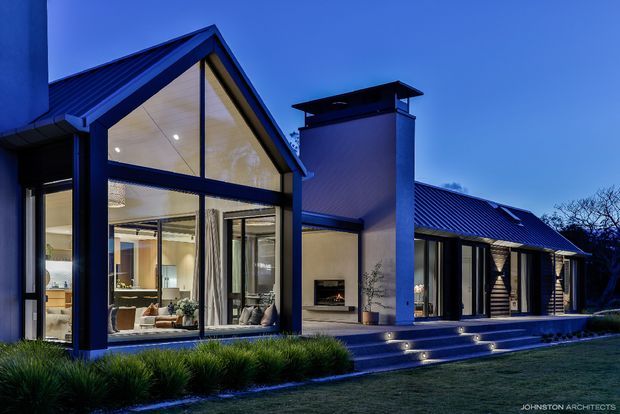 Dusk exterior view of a contemporary single-storey residence with a prominent steep gabled glass pavilion, dark metal framing, a long low pitched roof wing, and a tall chimney volume. Warm interior lighting reveals an open living area through the floor-to-ceiling glazing. Stepped entry terraces with integrated lights lead through landscaped planting to the home.