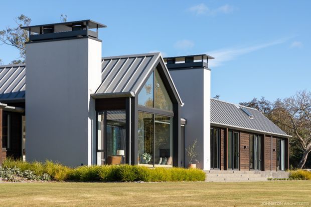 Exterior view of a contemporary single-storey residence with steeply pitched standing-seam metal roofs, two tall rectangular chimney-like roof structures, dark timber cladding, and extensive floor-to-ceiling glazing. The home sits within a landscaped lawn and garden, with a prominent glazed gable and strong geometric roof forms.
