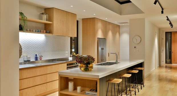 A warm, contemporary open-plan kitchen with extensive light timber cabinetry, a long white island with integrated sink and seating for several people, and a metallic mosaic-tile splashback. Recessed ceiling lighting and black track lights illuminate the kitchen, while a timber-floored hallway and entry area extend beyond it.