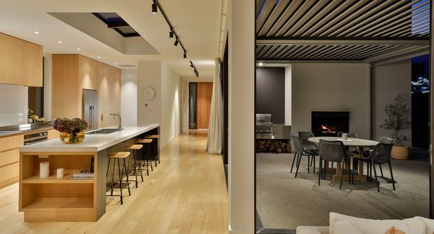 Split view of a refined contemporary home showing a light timber kitchen with a long white island and multiple stools on the left, opening toward a timber-floored hallway, and an adjoining outdoor or covered entertaining area on the right with dining furniture, a fireplace and a slatted overhead roof structure.
