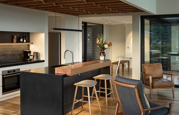 A contemporary open-plan kitchen and living area with a large dark island, timber breakfast bar, integrated appliances, and black-framed glazing. Warm timber ceiling battens and flooring contrast with pale cabinetry and dark stone-like surfaces. Timber stools, upholstered lounge chairs, and a leather armchair furnish the adjacent dining and living zone.