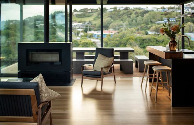 A refined open-plan living and kitchen area with expansive floor-to-ceiling glazing overlooking a leafy suburban landscape. The room features warm timber flooring, a dark built-in fireplace, mid-century-inspired lounge chairs, a streamlined kitchen island with timber cabinetry and a light benchtop, and bar stools. The space is bright, warm, and connected to the surrounding greenery.