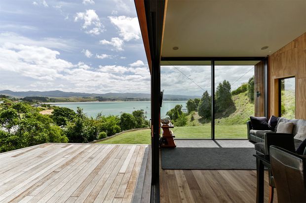 A bright contemporary living room opens directly onto a timber deck through expansive floor-to-ceiling glazing, framing panoramic views over a green landscaped site and calm coastal water. The interior features warm timber wall panelling and flooring, a dark-framed glazed wall, a charcoal rug, and dark upholstered seating. The deck and living space create a strong indoor-outdoor connection.