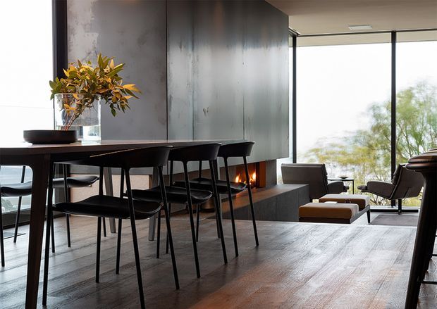 Contemporary open-plan interior combining a dining area and living space. A dark timber dining table with slim black chairs sits beside a large sculptural dark feature wall containing a linear fireplace. The room has broad timber flooring, floor-to-ceiling glazing, restrained furnishings, and views toward trees outside. The palette is dark, warm, and refined with strong natural light.