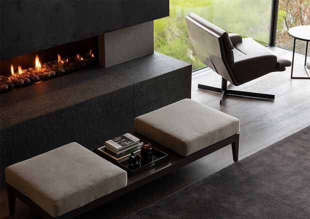 A refined contemporary living room with a linear gas fireplace set into a dark textured stone or concrete surround and broad hearth. Two upholstered ottomans sit around a low dark coffee table, while a modern lounge chair faces the fireplace beside full-height corner glazing. The room has a restrained neutral palette, strong geometric forms, and a warm firelit atmosphere.