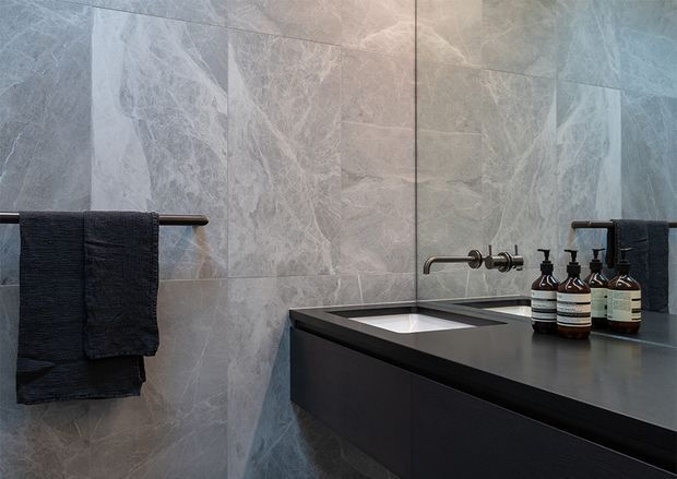 A sophisticated contemporary bathroom vanity area with large-format grey veined stone-look wall tiles, a dark integrated benchtop and floating vanity cabinetry, wall-mounted brushed metal tapware, a large mirror, and dark towels with neatly arranged toiletry bottles. The space has a moody, refined atmosphere and a clean minimalist fit-out.