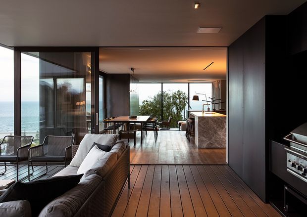 A contemporary open-plan coastal home interior with a dark, refined material palette. The foreground contains a living area with a large upholstered sectional sofa and lounge chairs, opening into a dining area and kitchen. Full-height glazing frames ocean views and brings in abundant natural light. Warm timber floorboards contrast with dark cabinetry and walls, while a stone-clad kitchen island and minimalist pendant lighting define the central space.
