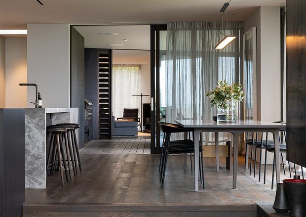 Contemporary open-plan dining and kitchen interior with a dark timber floor, stone kitchen island, black-framed glazed partition, and a dining table positioned beside floor-to-ceiling sheer curtains. A lounge area and stairway are visible beyond the open connection. The space uses restrained dark neutrals, warm timber, pale stone, and sculptural pendant lighting.