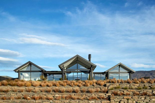 A low-profile contemporary residential building composed of multiple glazed gabled pavilions, set into a terraced rocky landscape with ornamental grasses. The dark metal-framed glazing and muted roof cladding contrast with extensive stone retaining walls, while distant hills and a blue sky form the background.