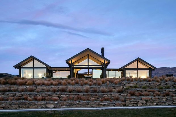 A low-slung contemporary single-storey house viewed from the exterior at dusk. The composition features three prominent gabled glazed volumes linked by a lower central section, dark structural framing, extensive floor-to-ceiling glazing, and warm interior lighting. Layered stone retaining walls and sparse tussock planting form the foreground, with a dry mountainous landscape visible beyond.